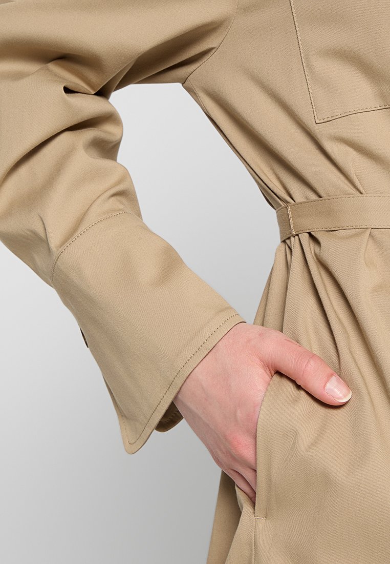 Beżowy płaszcz trench z paskiem w talii, z długimi, lekko rozszerzającymi się rękawami oraz boczną kieszenią, wykonany z gładkiego materiału.