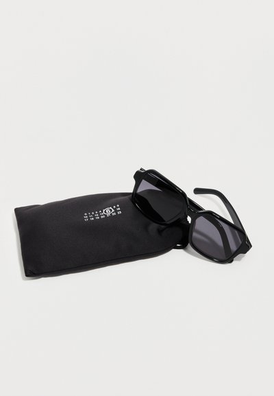 Μαύρα ορθογώνια γυαλιά ηλίου που ακουμπούν σε μαύρη μαλακή θήκη με έναν λευκό κύκλο που περιέχει τον αριθμό 6 και μικρούς αριθμούς τοποθετημένους από πάνω.