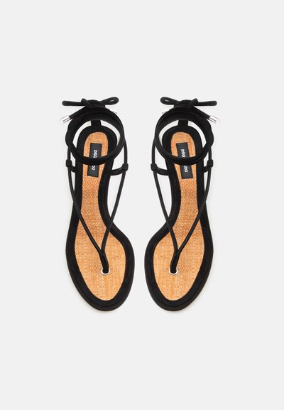 Sandales plates noires à brides avec semelles tressées couleur tan et liens à nouer autour de la cheville, présentées côte à côte sur un fond blanc.