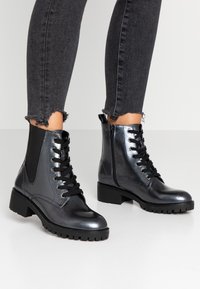 Botas de tobillo de charol negro con suela de tacos, que presentan cordones en la parte frontal, cremalleras laterales y paneles texturizados en los laterales.