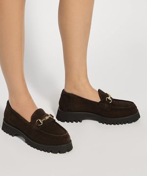 Jambes portant des mocassins en daim marron foncé avec des semelles épaisses noires et un détail en métal doré en forme de mors sur un fond clair uni.