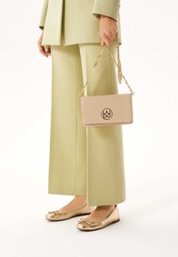 Béžová crossbody taška so zlatým reťazcom, ktorá má okrúhle logo. Nosená s svetlozelenými nohavicami a kovovými zlatými balerínkami.