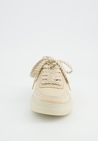 Baskets beige avec une tige en cuir lisse, une semelle en caoutchouc épaisse et des lacets en corde torsadée texturée. Détails en métal doré aux extrémités des lacets.