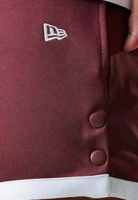 Pantalones cortos deportivos burdeos con tejido texturizado, notable ribete blanco y dos botones oscuros en el lado. Presenta un logo bordado arriba.