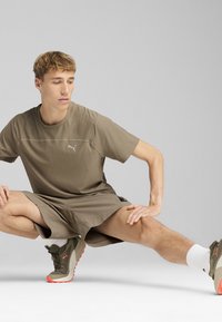 Puma aktiven komplet, ki vključuje olivno zeleni kratki rokav in ujemajoče se hlače, narejen iz lahkega materiala, skupaj s sivo-oranžnimi supergami.