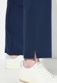 Marinblå byxor med sidoflikar vid fållen, tillverkade av ett slätt tyg. Kombinerade med vita sneakers med en ljusgrå accent.