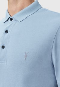 Polo shirt di colore azzurro chiaro con tessuto testurizzato, tre bottoni neri e un piccolo design di teschio ricamato sul lato sinistro del petto.