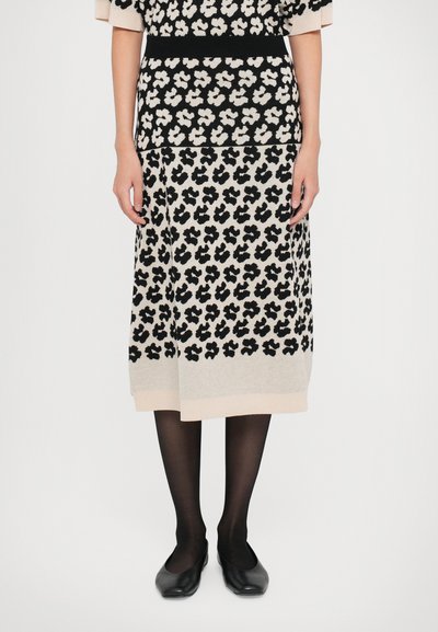Vestido de punto con patrón floral negro, fondo crema, mangas cortas, cinturilla negra gruesa y detalle en el dobladillo. Combinado con zapatos planos negros y medias.