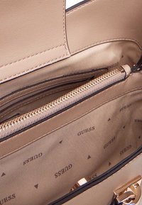 Interno di una borsa in finta pelle beige, con una tasca con zip, rivestimento beige liscio con motivo logo ripetuto e accenti in trim scuro.
