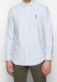 Ljusblå och vit randig skjorta med lång ärm och knappkrage, med en liten marinblå logotyp på bröstet och manschetter med knappar.