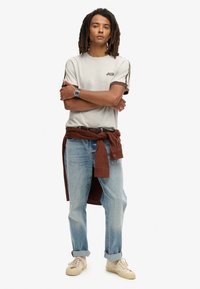 Lysegrå kortærmet t-shirt med logo, kombineret med lyseblå jeans. Brun skjorte bundet om taljen og cremede sneakers med snørebånd.