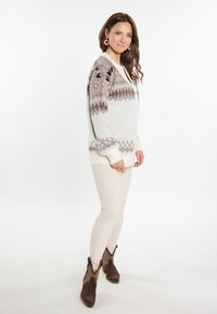 Cárdigan gris claro con un patrón geométrico en marrón y crema; textura suave, con botones al frente; combinado con pantalones ajustados beige y botas de tobillo marrones.
