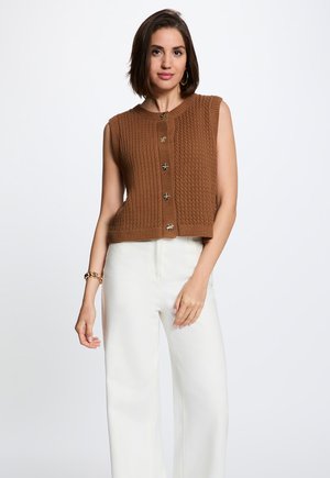 Donna che indossa un gilet marrone senza maniche lavorato a maglia con bottoni decorativi e pantaloni bianchi a gamba larga, in piedi davanti a uno sfondo semplice.