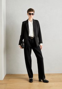 Blazer nero su una camicia bianca, abbinato a pantaloni neri larghi e scarpe con tacco spesso. Inclusi occhiali da sole neri e una piccola borsa nera.
