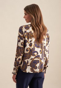 Blusa con bottoni dal motivo floreale in blu navy e giallo dorato su uno sfondo crema. Maniche lunghe con dettagli ai polsi.
