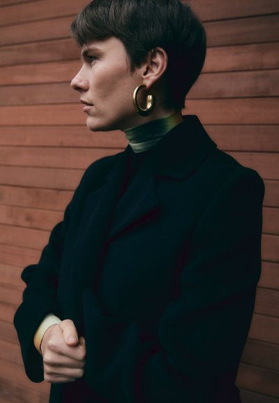 Czarny płaszcz z kołnierzem, jasna zielona golfka i złote kolczyki w kształcie kół. Modelka stoi na tle drewnianym z dłońmi złożonymi.
