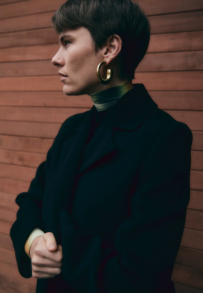 Svart kappa med krage, ljusgrön polotröja och guldörhängen. Modellen står mot en träbakgrund med händerna sammanfogade.