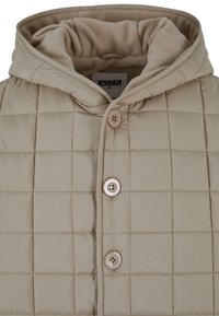Beige quiltet pufferjakke med hætte, der har et blødt indvendigt for og tre store knapper foran til lukning.