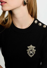 Maglione nero a costine con bottoni dorati sulla spalla e una toppa decorativa ricamata con una corona ornata e la lettera "M."