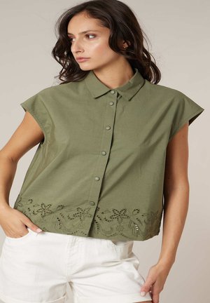 Femme portant un haut sans manches boutonné couleur vert olive avec col et broderie florale le long de l'ourlet, associé à un pantalon blanc.