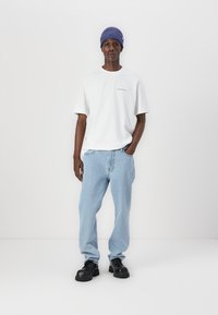 Tricou alb cu mânecă scurtă, blugi deschiși la culoare și cizme negre cu șireturi. Modelul poartă o căciulă albastră cuべ și stă pe un fundal alb.