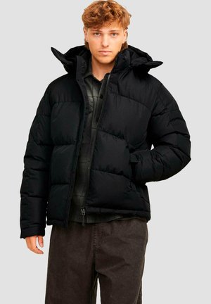 Young man with light brown hair wearing an unzipped black puffer jacket over a dark plaid shirt and dark pants against a plain background.