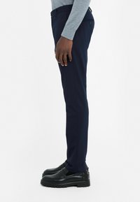 Marineblauwe broek met een slim fit, voorzien van een soepele textuur en twee zijzakken, gecombineerd met zwarte instap schoenen op een witte achtergrond.