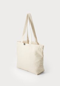 Borsa tote in tela beige con due manici lunghi, forma quadrata e un piccolo tag con logo sul fronte. Texture liscia, design minimalista.