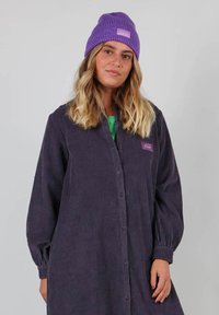 Robe à boutons en velours côtelé violet foncé avec des manches longues et une texture côtelée, associée à un bonnet violet assorti comportant un patch logo.