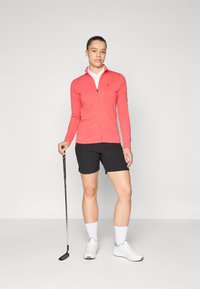 Korálová zipová bunda s dlouhými rukávy, spárovaná s černými šortkami. Pod ní bílá košile s límečkem a bílé sportovní boty. Golfová hůl držená v pravé ruce.