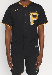 Uomo che indossa una maglia sportiva nera con bottoni e la lettera "P" gialla sul petto, con colletto e maniche bordati di giallo, abbinata a pantaloni corti Nike neri.
