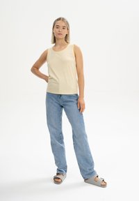 Lichtgele mouwloze top met horizontale strepen, gecombineerd met lichtblauwe rechte jeans. Model draagt grijze sandaal met een gesp.