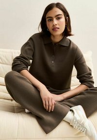 Pull polo en tricot marron foncé avec trois boutons, associé à un pantalon assorti. Le modèle porte des baskets blanches et est assis sur un fond texturé crème.
