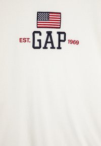 Sudadera de felpa blanca con bandera americana bordada, "GAP" en azul marino y "EST. 1969" en rojo, centrado en el pecho. Textura suave.