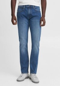 Jeans de denim azul con un corte recto, que cuentan con cinco bolsillos, un cierre de botón y cremallera, y detalles de costura visibles. Combinados con zapatillas grises.