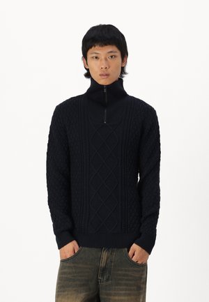 Pull en maille bleu marine avec un col montant et une demi-fermeture éclair, présentant des motifs texturés de câbles et de cercles, ainsi que des poignets et un ourlet côtelés.