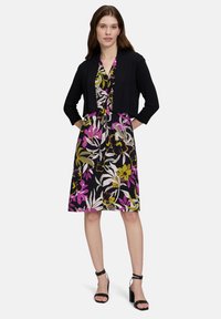 Jongeman in een bloemenjurk met een zwarte cardigan en zwarte hakken met bandjes, die zelfverzekerd staat tegen een effen witte achtergrond.