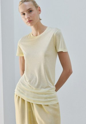 Kvinde med blondt hår iført en bleg gul T-shirt med korte ærmer og matchende løstsiddende bukser mod en ensfarvet lys baggrund.