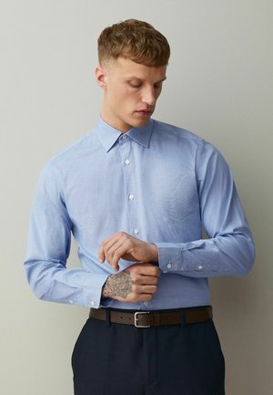 Young man with short curly hair wearing a light blue buttoned shirt and dark pants adjusts his cuff, showing a detailed geometric hand tattoo.