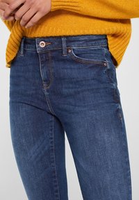 Blue denim jeans with a slim fit, featuring copper hardware, contrast stitching, and front pockets. The fabric has a smooth, slightly worn texture.