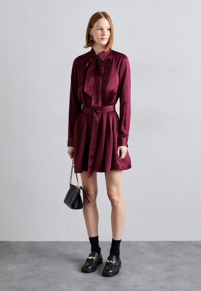 Vestido de satén burdeos con pliegues, cuello rizado y lazo. Combinado con mocasines negros y un pequeño bolso negro.