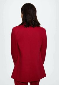 Femme avec des cheveux bruns mi-longs portant un blazer rouge ajusté et un pantalon assorti, vue de dos sur un fond uni.