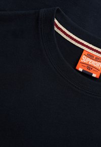 T-shirt bleu marine à col rond avec un col intérieur rayé rouge et blanc et une étiquette de marque Superdry orange cousue à l'intérieur du col.