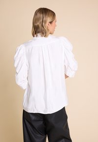 Hvit bluse med puffede ermer og plisserte detaljer bak, laget av et glatt stoff. Båret med svarte, løst sittende bukser.