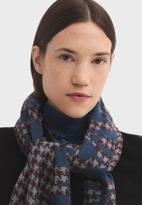 Écharpe tissée avec un motif pied-de-poule en bleu et bordeaux, en tissu tricoté texturé, enroulée autour du cou.