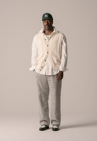 Vesta tricotată de culoare crem, cu butoane din lemn, purtată peste o cămașă albă. Pantaloni gri cu model și pantofi alb-negru. Accent de culoare verde de pe șapcă.