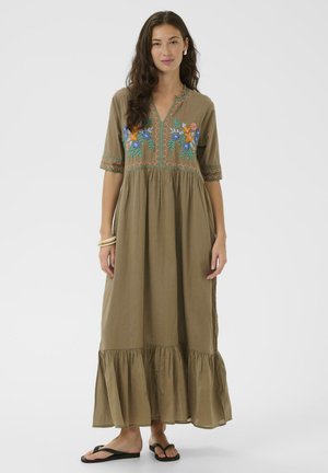 Femme portant une longue robe vert olive avec une broderie florale colorée sur la poitrine et les manches, associée à des tongs noires et des bracelets beiges.