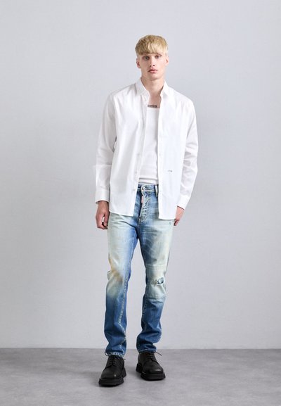 Giovane uomo con capelli biondi corti che indossa una camicia bianca aperta sopra una canotta bianca, jeans blu sbiaditi e strappati, e scarpe nere, in piedi contro un muro semplice.