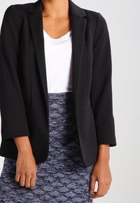 Černý blazer s texturovanou tkaninou, přizpůsobený střih, klopové límce a dvě přední kapsy, oblečený přes bílé tričko a vzorovanou sukni.