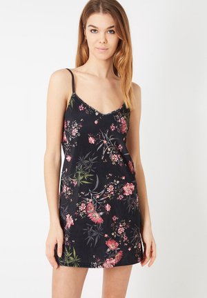 Mujer joven con un vestido negro sin mangas con flores y tirantes finos, de pie frente a un fondo blanco liso.